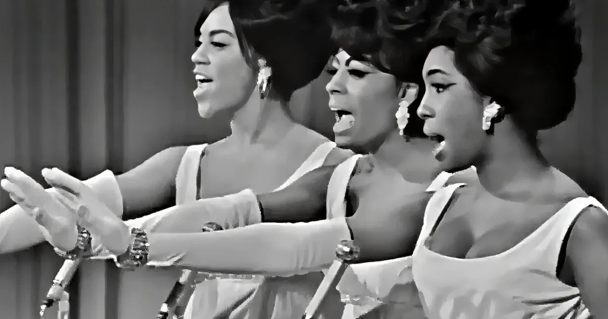 Three women in white dresses sing passionately, their arms extended, wearing earrings and bracelets.