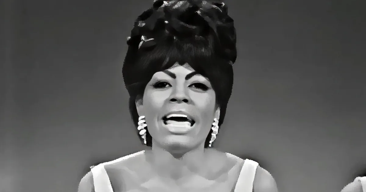 A woman with a large, styled hairdo and white earrings is singing.