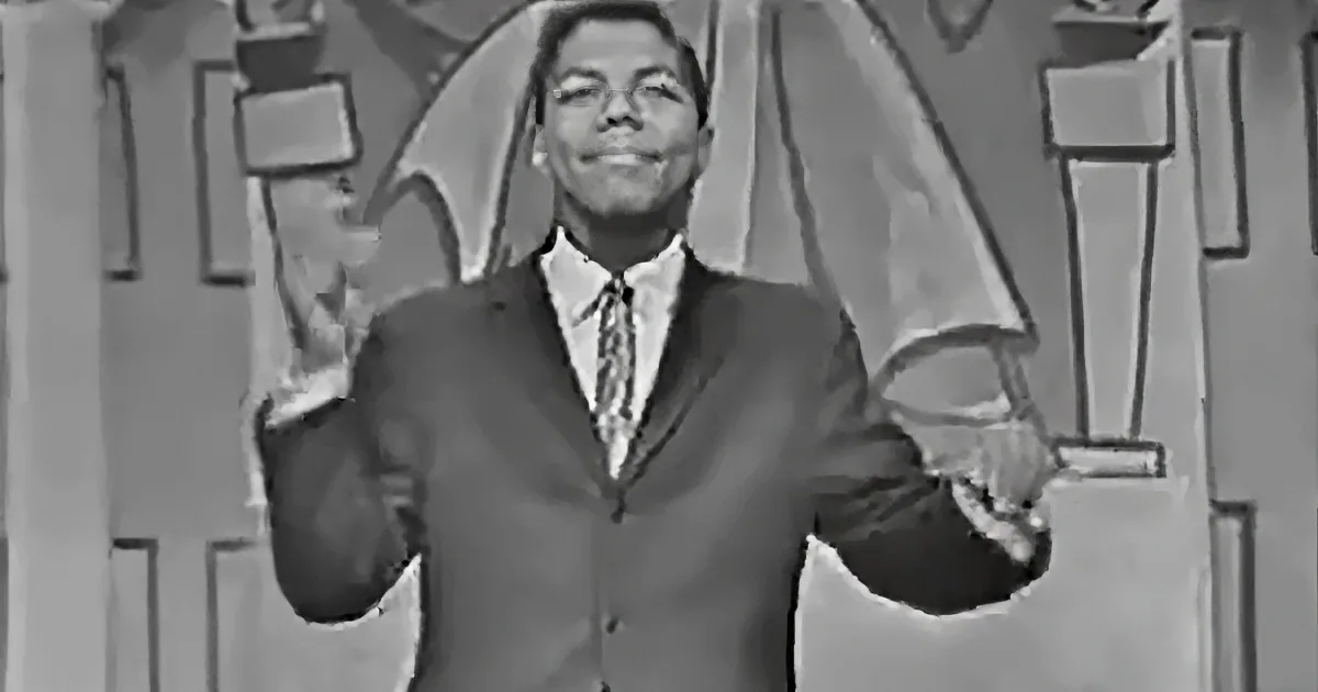 A man in a suit and tie smiles, raising his hands in a celebratory gesture.