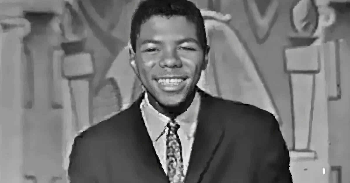 A smiling man in a suit and tie, wearing a patterned tie, stands in front of a decorative background.