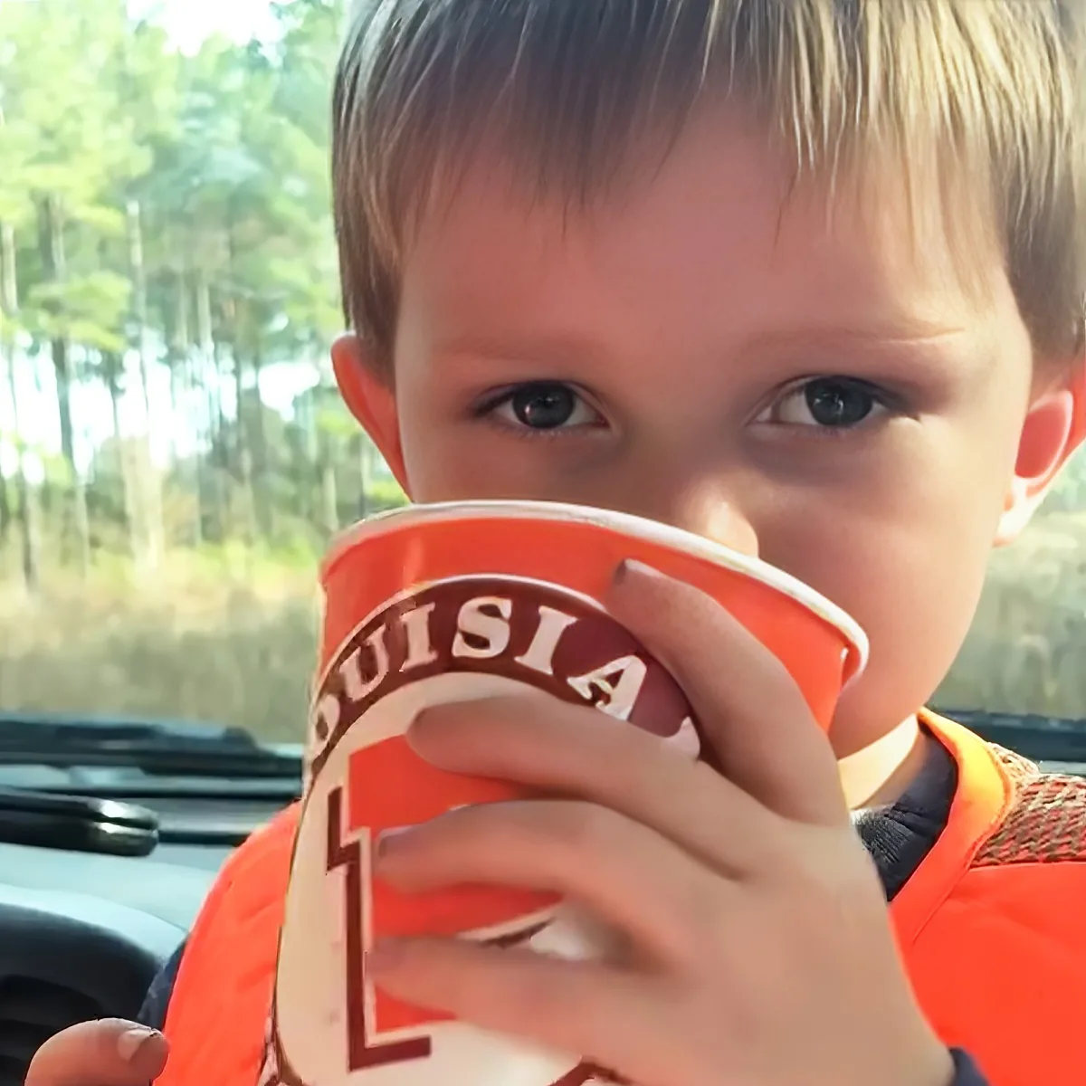 A young boy in an orange shirt holds a red cup with "Louisiana" on it, drinking from it.