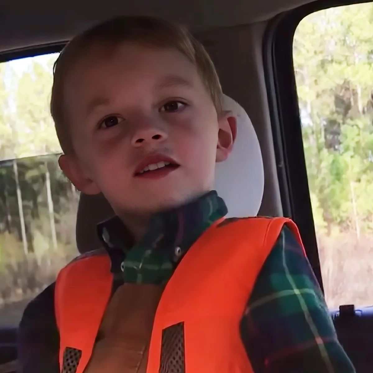 A young boy wearing an orange safety vest sits in a car, looking forward with a neutral expression.