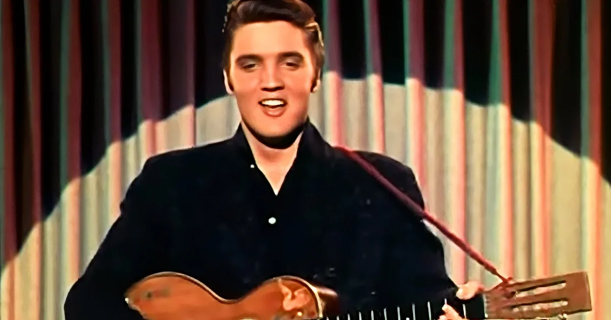 Elvis Presley in a black suit plays a guitar on stage with a colorful backdrop.