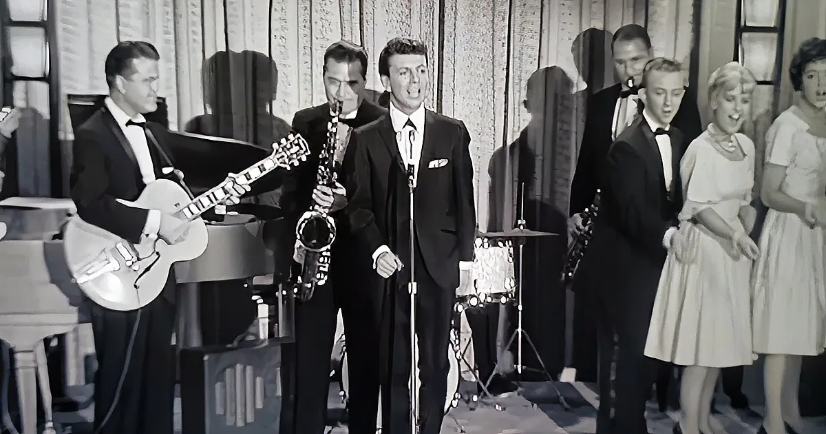 A man in a suit sings into a microphone, accompanied by musicians and a band.