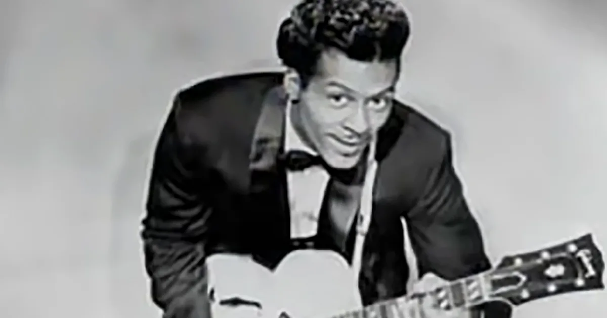 A man in a tuxedo with a bow tie and a guitar, smiling and looking to the side.