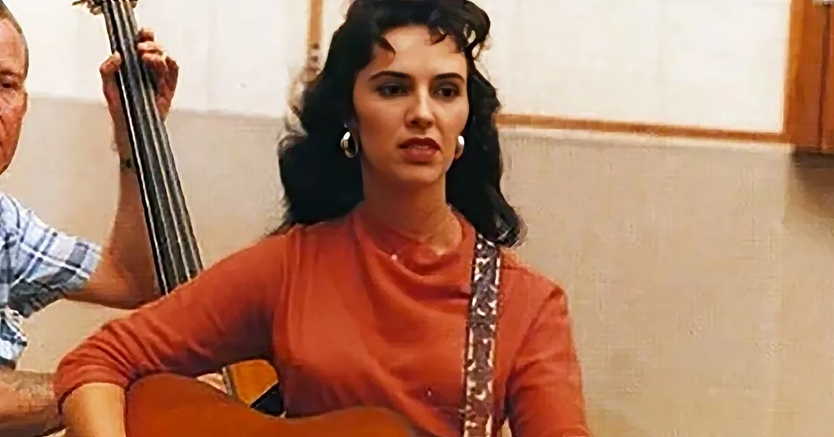A woman in an orange shirt plays a guitar while a man in a plaid shirt plays a bass.