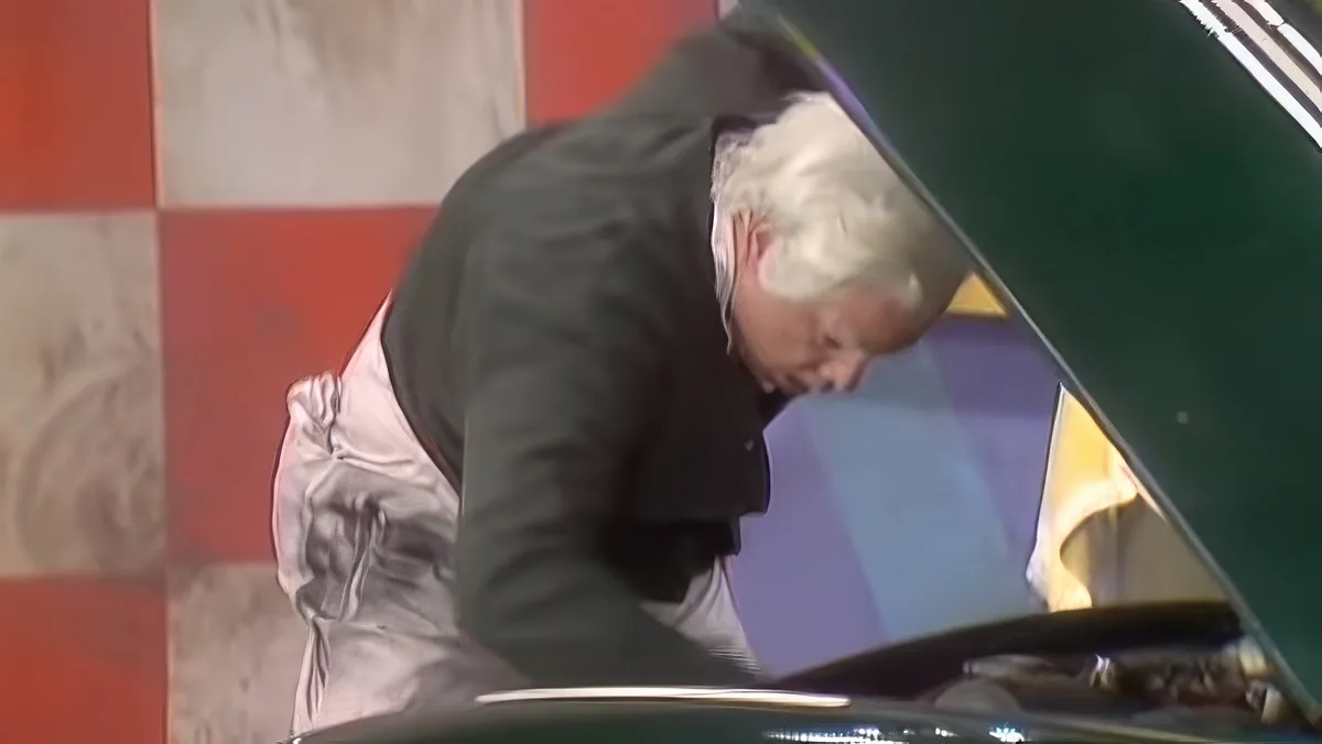 An older man in a dark jacket leans over a green car, inspecting or working on it.