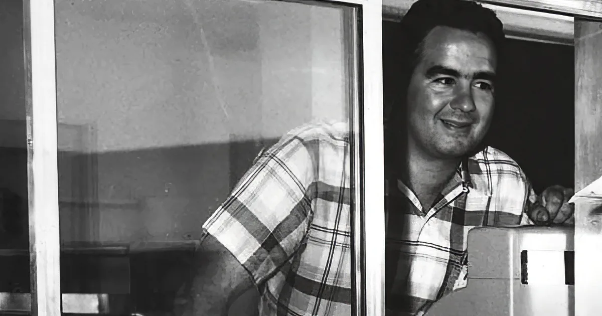 A man in a plaid shirt smiles near a window, with a blurred background and a reflection visible.
