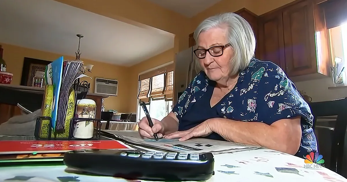 An elderly woman with gray hair and glasses is writing at a table with a calculator and papers.