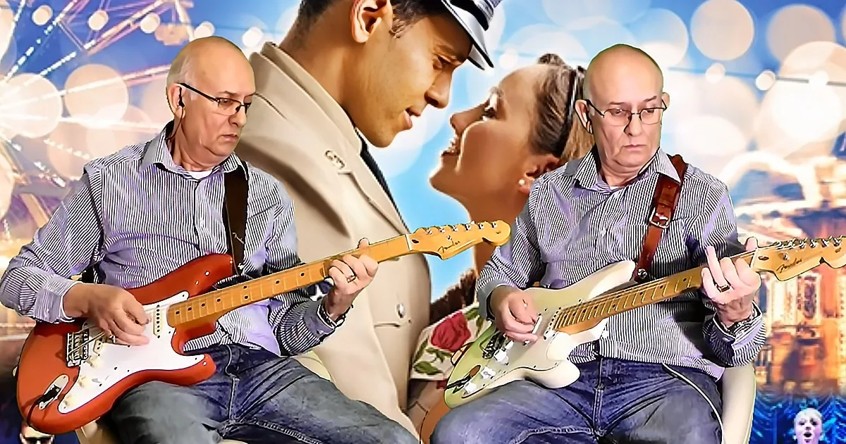 A man in a beige coat plays guitar with two others, one in a striped shirt, against a colorful, bokeh background.