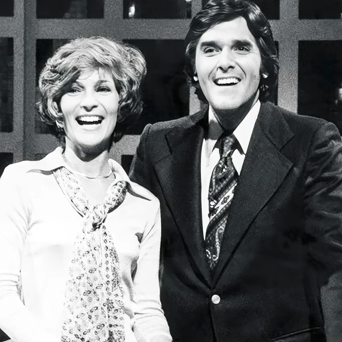 A smiling woman in a patterned blouse and a man in a suit and tie pose together.