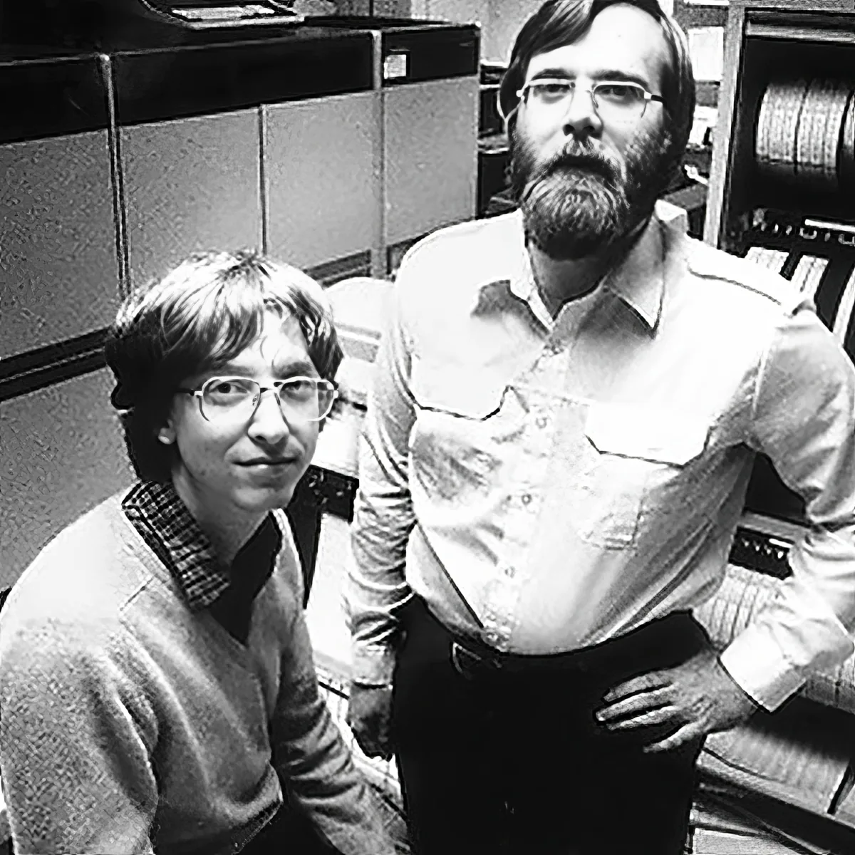 Two individuals in a workspace, one seated and one standing, both wearing glasses, with a vintage office setting in the...