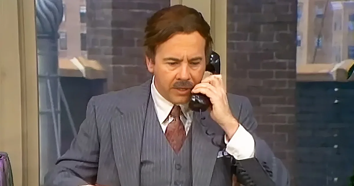 A man in a pinstripe suit and tie is holding a phone to his ear, appearing to be in a conversation.