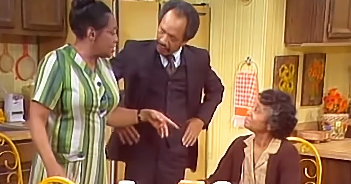 A man in a suit gestures while talking to a woman in a striped dress in a kitchen setting.