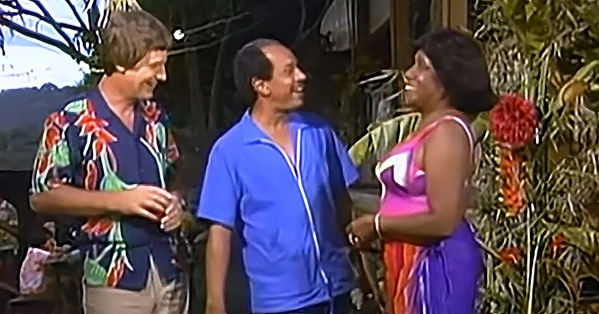 Three people in colorful outfits are smiling and talking outdoors near tropical plants.