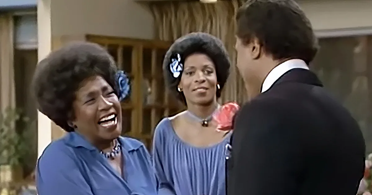 A woman in a blue shirt laughs while another woman in a blue dress and a man in a suit look on.