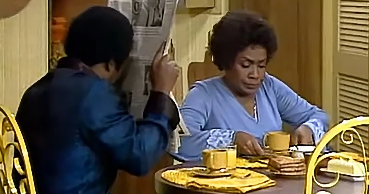 A man in a blue shirt is reading a newspaper while a woman in a blue top sits at a table with a cup of coffee.