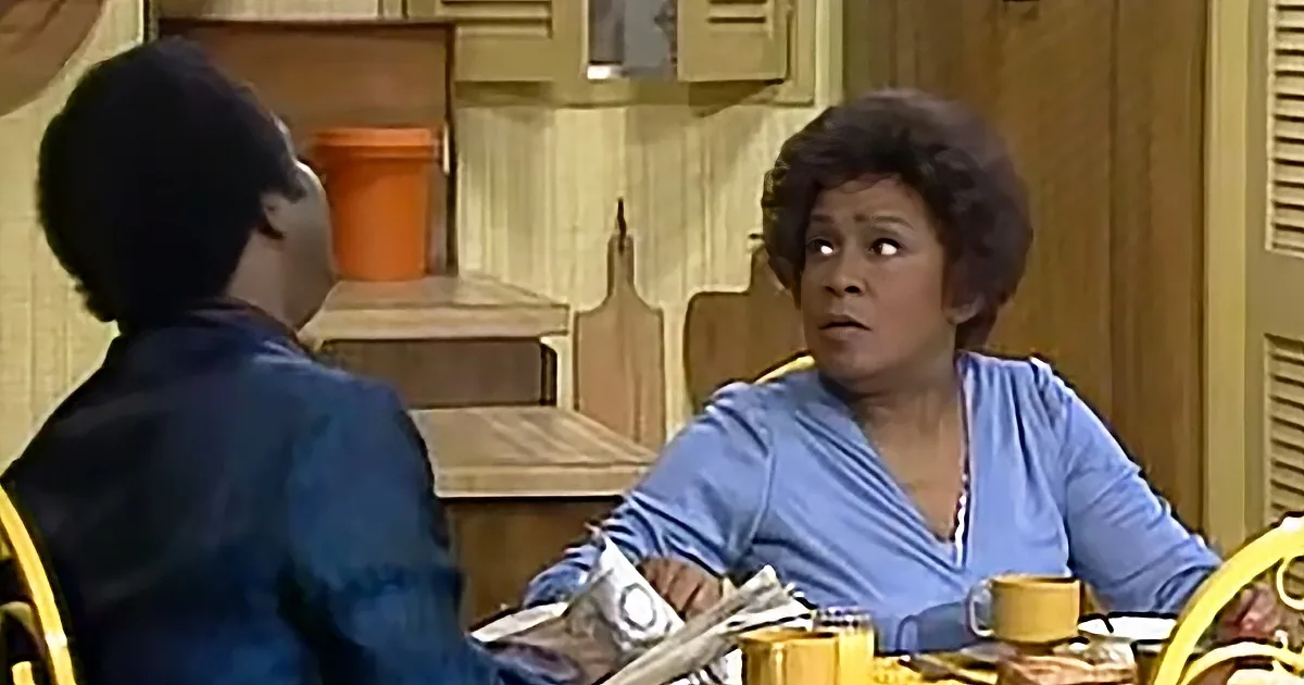 A woman in a blue blouse sits at a table, looking concerned, with a man in a dark shirt opposite her.