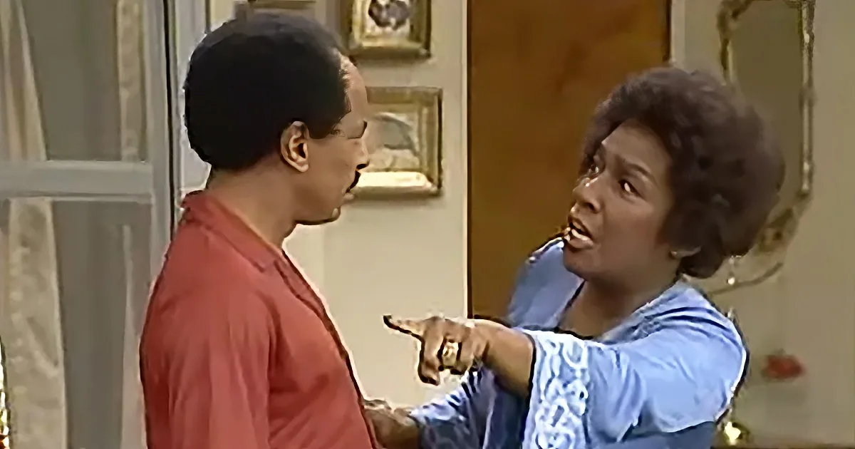 A woman in a blue dress points at a man in a red shirt in a room with framed pictures.