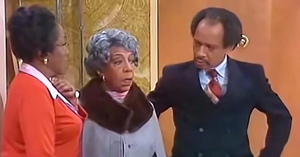 A woman in a gray coat and scarf is speaking, flanked by a man in a suit and a woman in an orange sweater.