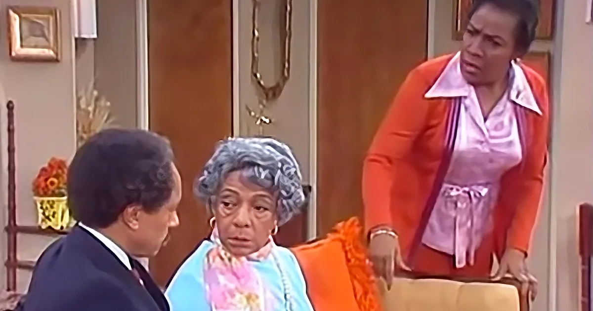 A woman in an orange outfit stands beside a seated woman in a blue dress, while a man in a suit looks on.