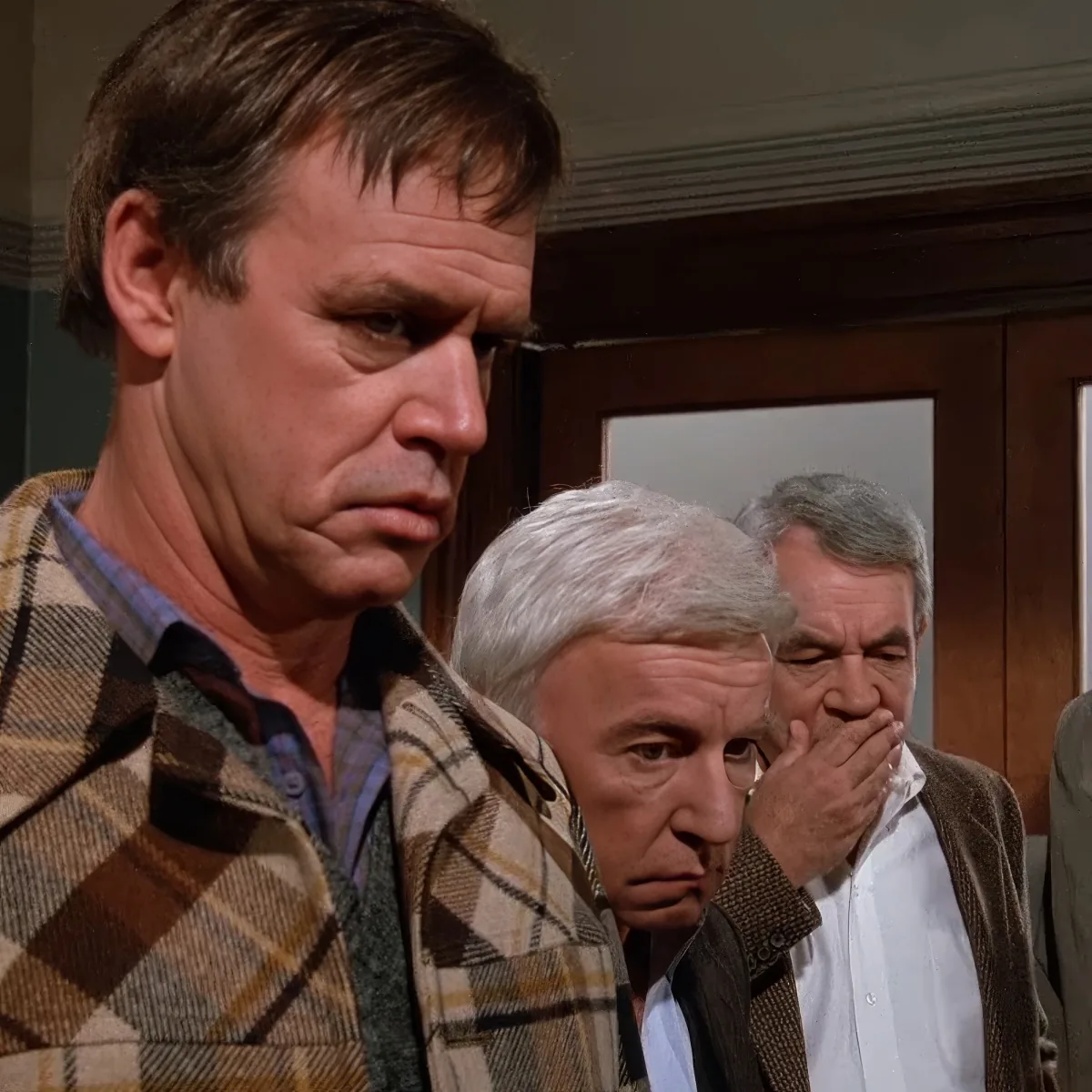 A man in a plaid jacket looks concerned, with two other men behind him, one covering his mouth.