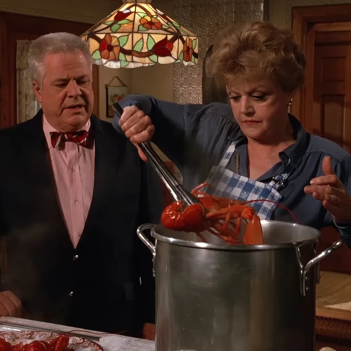 Woman in apron holding tongs over lobster in pot, man in suit looks on.