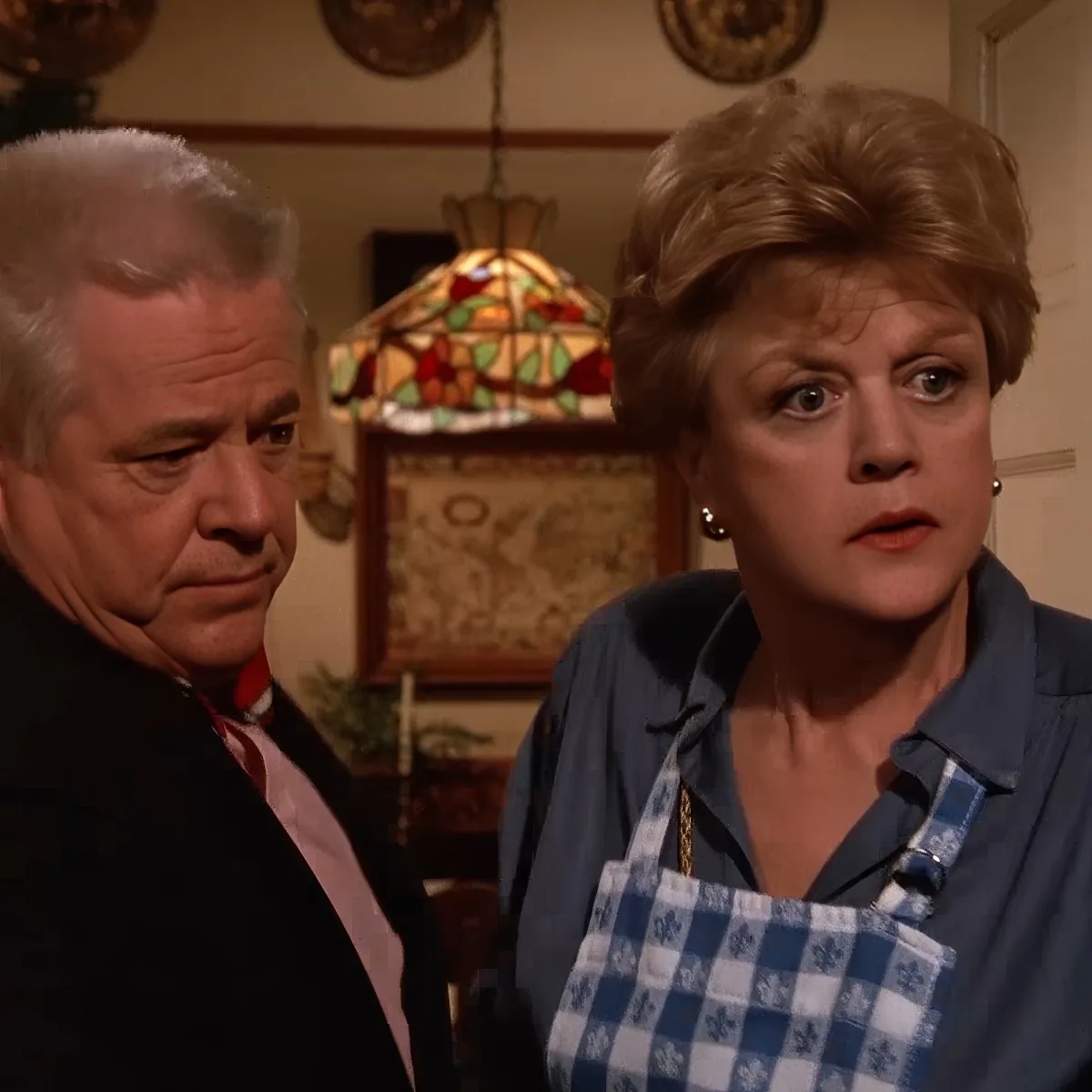 A woman in a blue shirt and checkered apron looks surprised, while a man in a black shirt and red tie looks concerned.