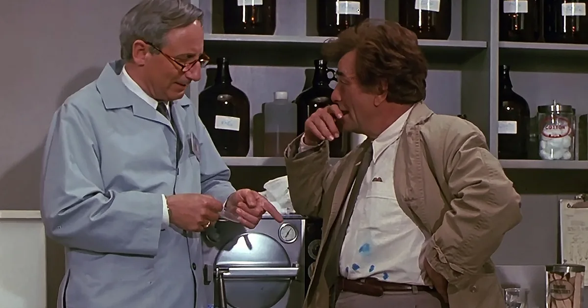 A man in a lab coat points at another man in a tan jacket, both in a laboratory setting with shelves of bottles.
