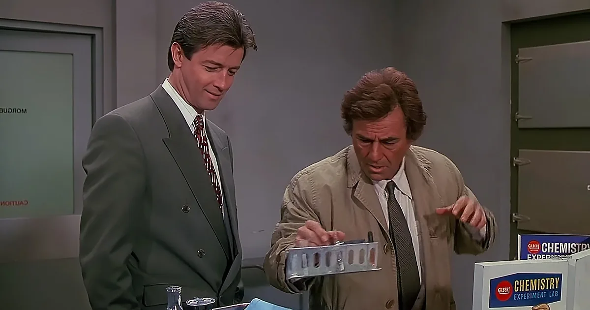 A man in a tan jacket points at a metal object while another man in a suit looks on.