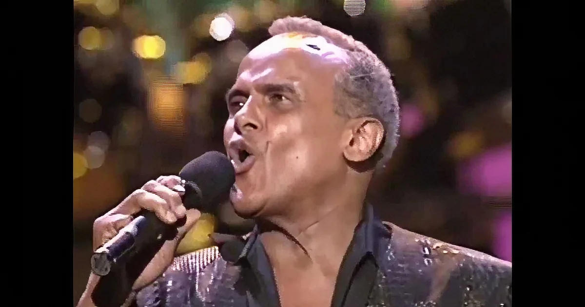 A man in a black outfit sings into a microphone on stage, illuminated by stage lights.