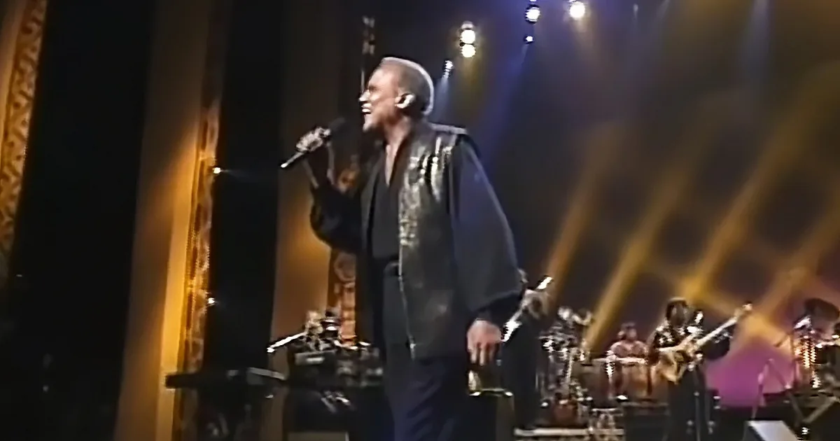 A man in a black outfit sings into a microphone on stage with a band in the background.