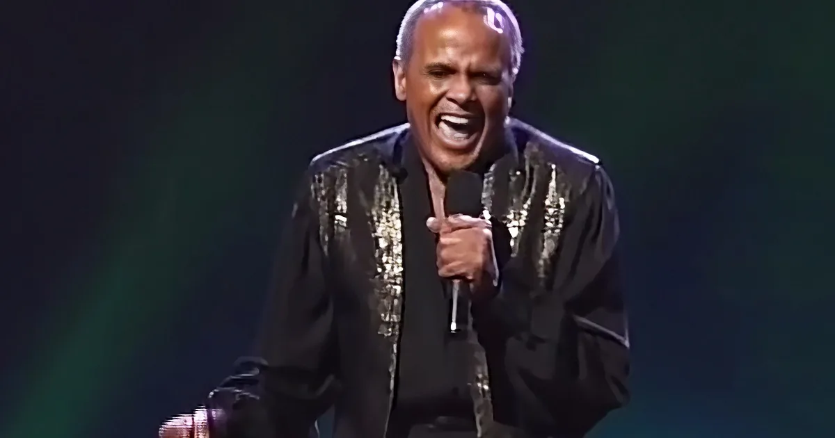 A man in a black sequined jacket sings passionately into a microphone on stage.