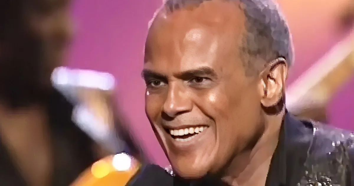 A smiling man with short hair and a black jacket, holding a microphone, in a blurred background.