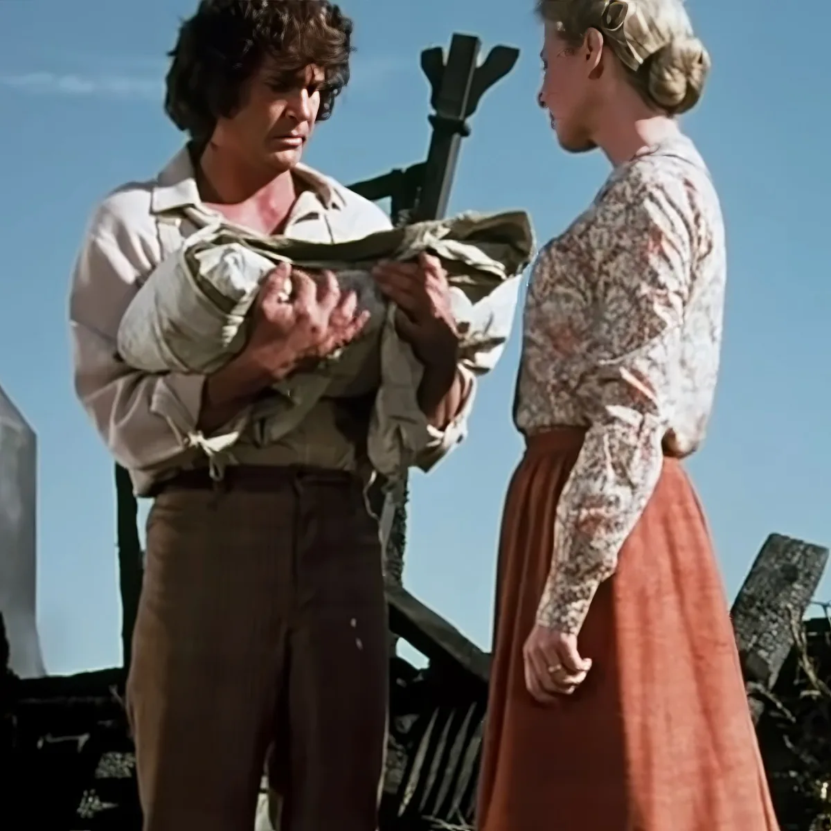 A man in period clothing holds a bundle, facing a woman in a floral top and skirt, against a clear sky backdrop.