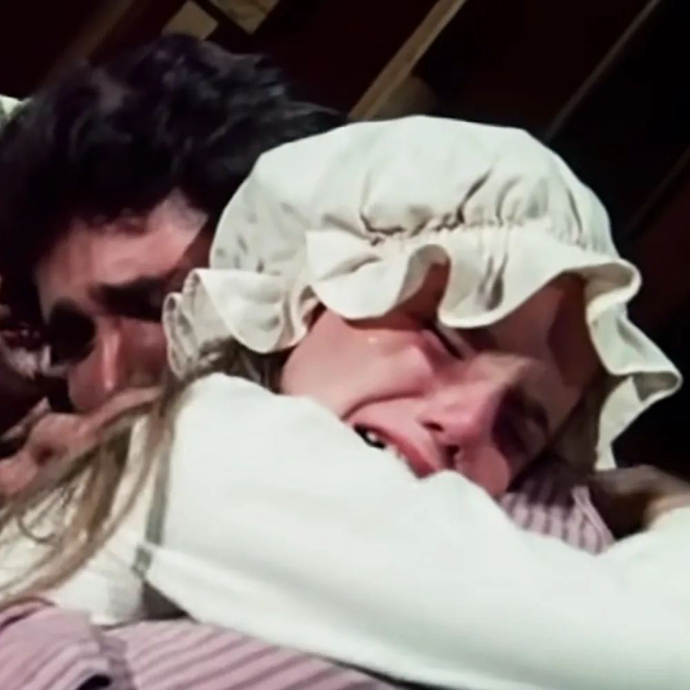 A person in a white bonnet and striped dress is lying down, with another person leaning over them.