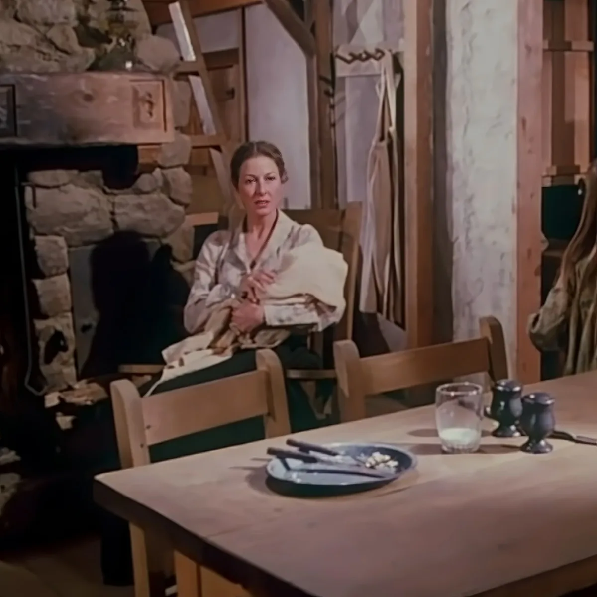A woman in a robe sits at a rustic table with a plate, glass, and condiments, in a stone-walled room.