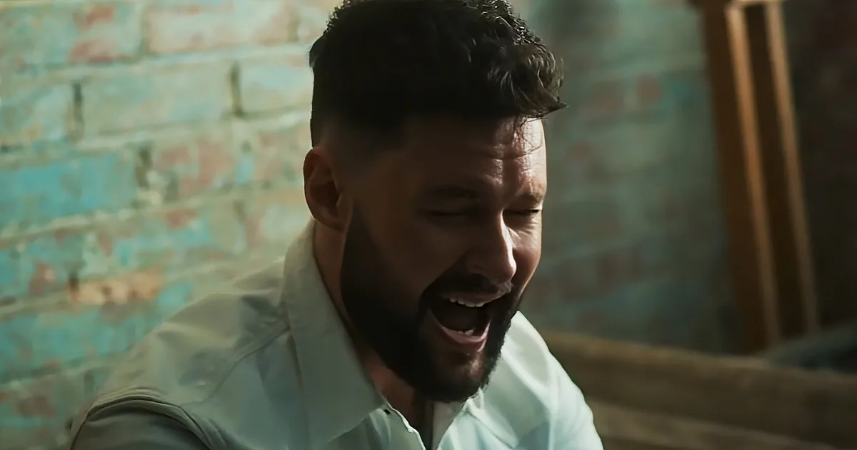 A man with a beard and short hair, wearing a light blue shirt, is laughing or speaking with his eyes closed.