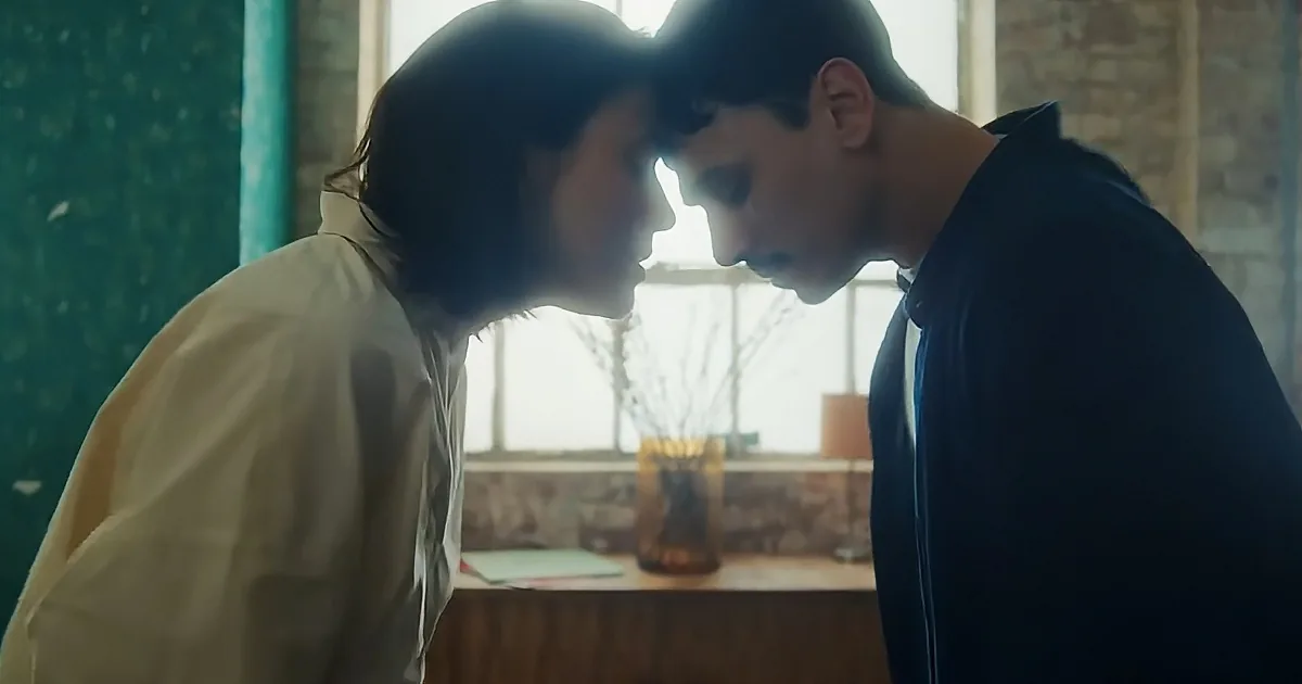 Two people are close, facing each other, with a window and vase in the background.
