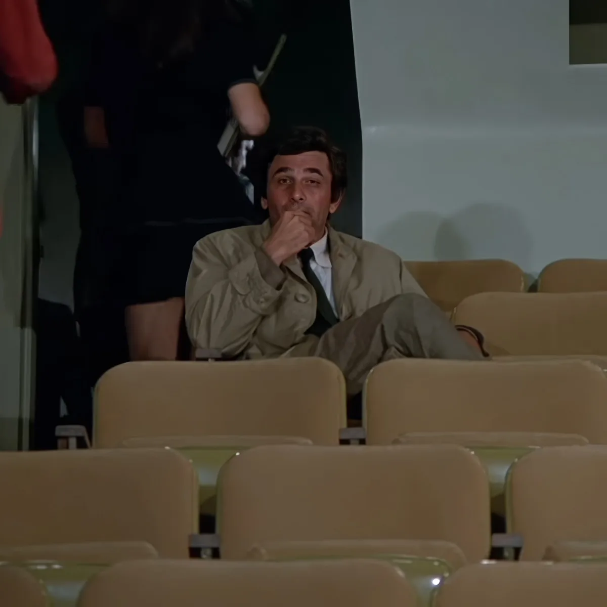 A man in a beige jacket sits in an empty auditorium, holding his hand to his mouth, appearing contemplative.