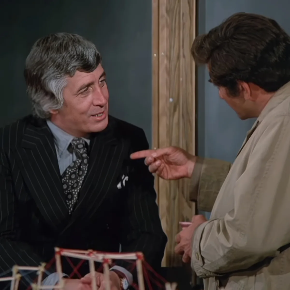 A man in a suit is engaged in a conversation with another person pointing at him, with a model structure in the foreground.