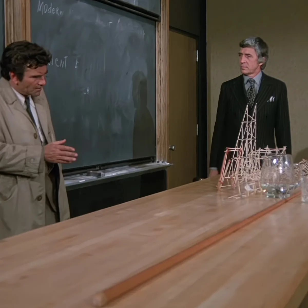 A man in a trench coat gestures near a chalkboard while another man in a suit stands beside a model structure on a table.