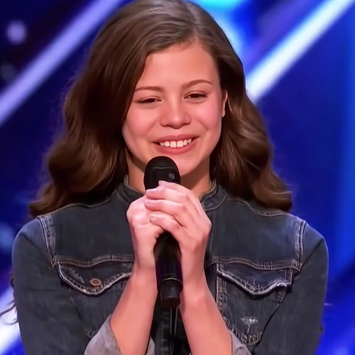 A person in a denim jacket holds a microphone, smiling with a blue stage background.
