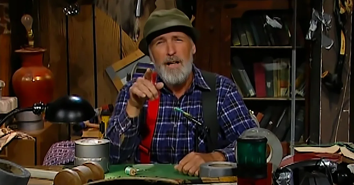 A man in a plaid shirt and hat sits at a cluttered workbench, gesturing with his right hand.