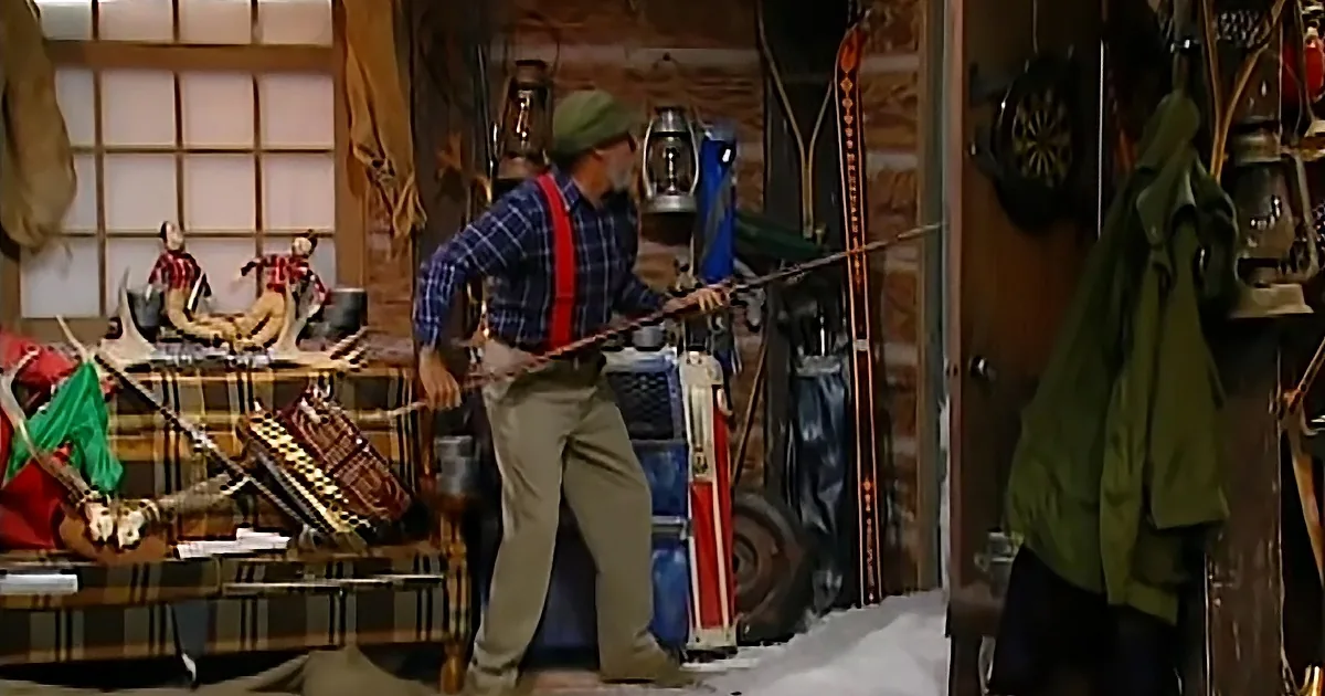 Man in plaid shirt and suspenders holds a bow and arrow in a cluttered room with hunting gear.