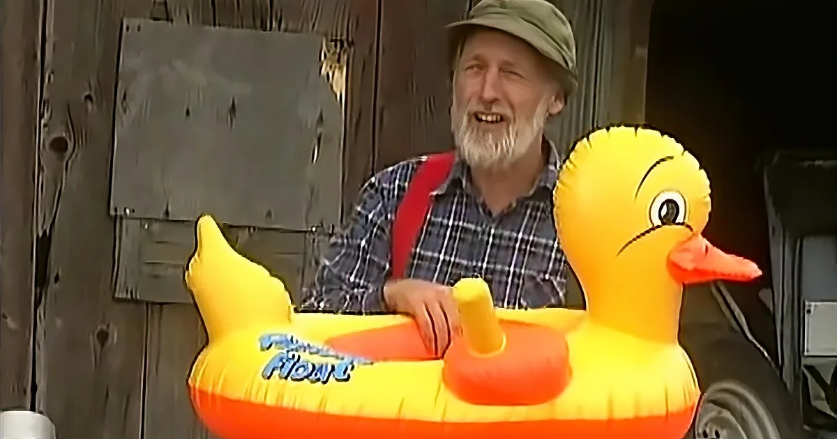 An older man in a plaid shirt and hat holds a large inflatable duck float.