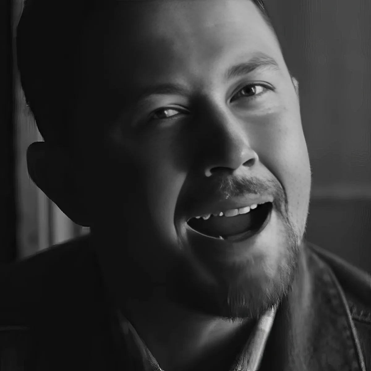 A man with a beard and mustache, wearing a dark jacket, smiles warmly in a black-and-white portrait.