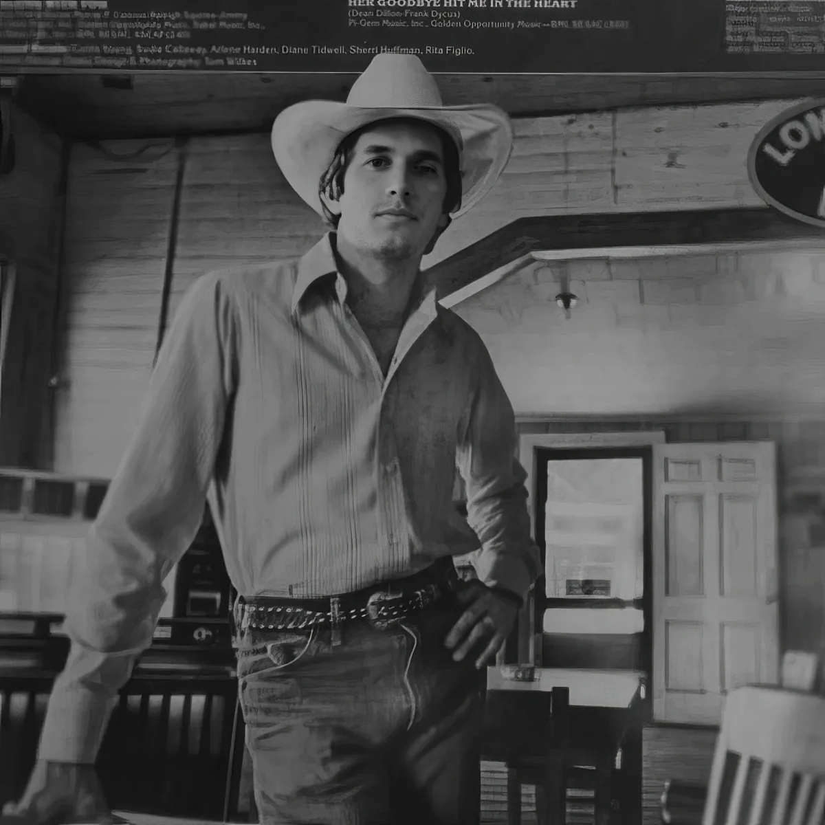 A man in a cowboy hat and western attire stands confidently in a rustic setting.