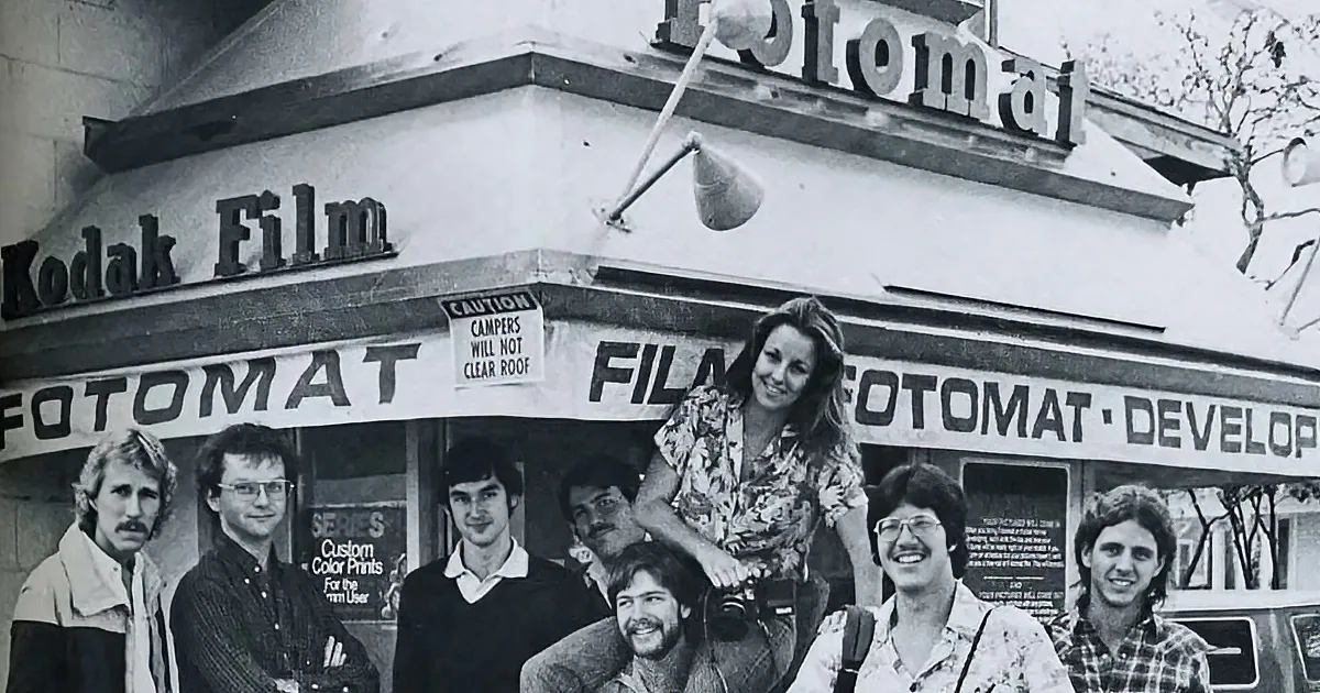 A group of people stands in front of a Fotomat store with a Kodak Film sign above.