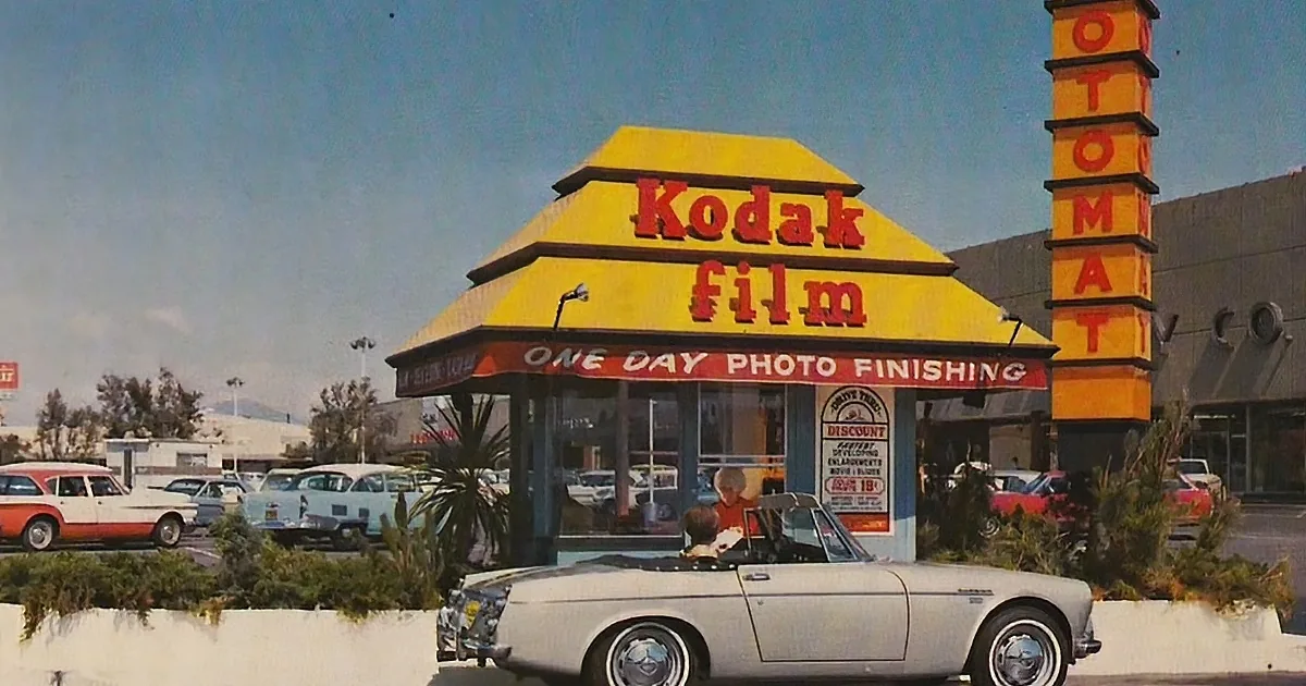 A vintage Kodak film booth with a yellow and red sign, surrounded by classic cars and palm trees.